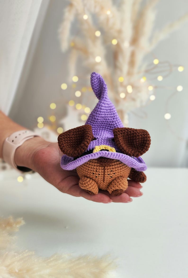 May include: A small crocheted brown dog wearing a purple witch hat with a yellow brim. The dog is sitting in the palm of a hand.