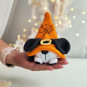 May include: A crocheted stuffed dog wearing an orange witch hat with a black spiderweb design. The dog has black ears, a white body, and black eyes.