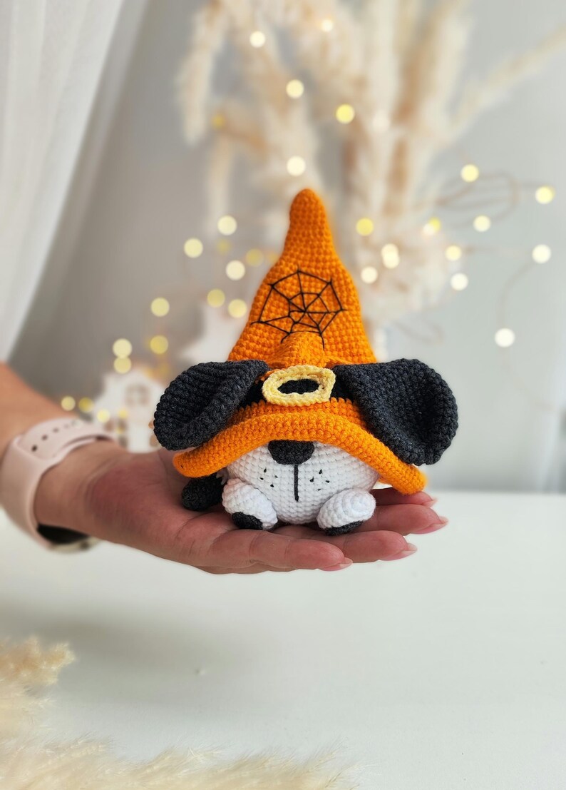May include: A crocheted stuffed dog wearing an orange witch hat with a black spiderweb design. The dog has black ears, a white body, and black eyes.