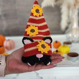 May include: A handmade crochet gnome with a red and cream striped hat adorned with yellow sunflower accents. The gnome has black ears and a black body. The gnome is being held in a hand.