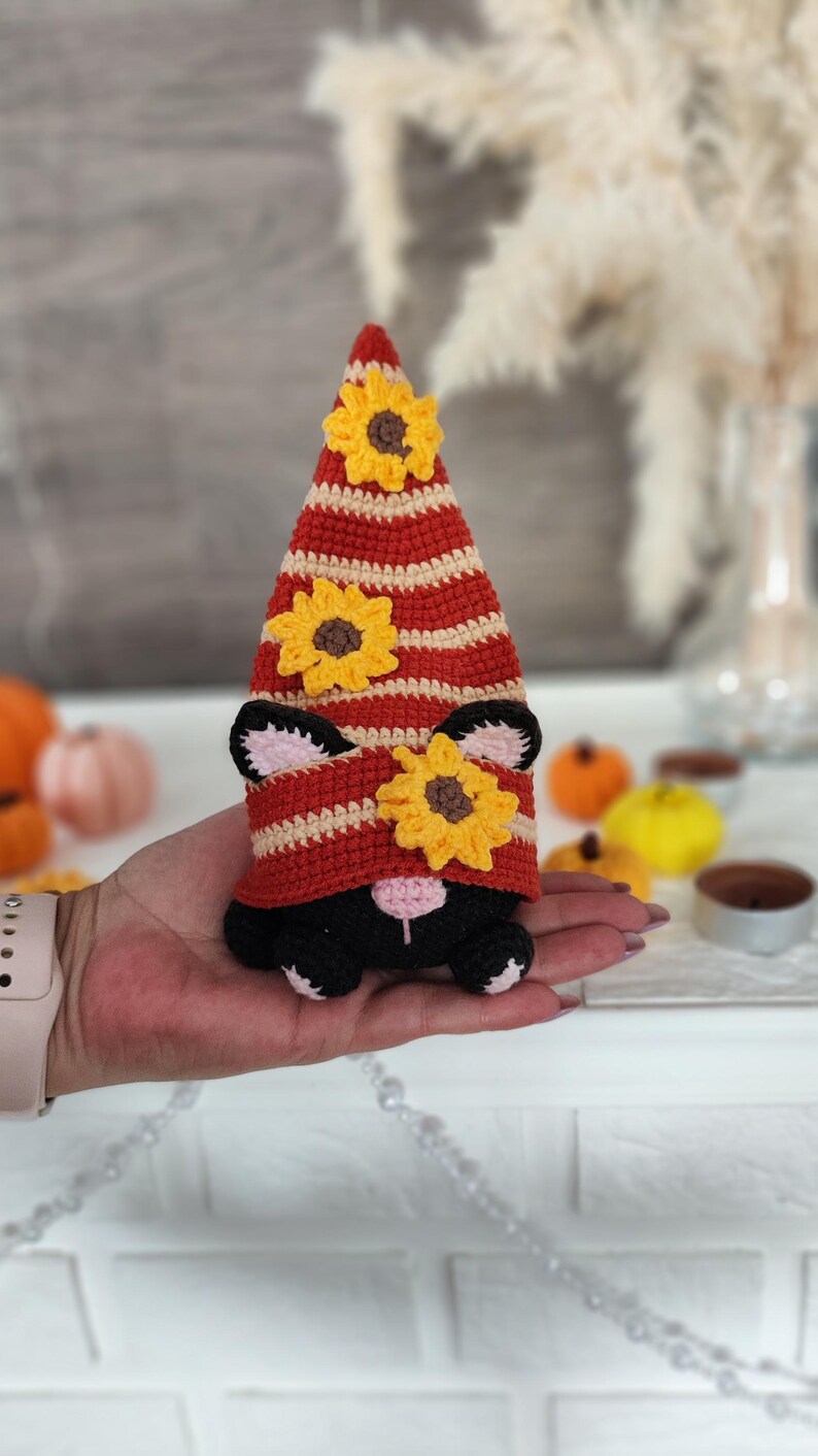 May include: A handmade crochet gnome with a red and cream striped hat adorned with yellow sunflower accents. The gnome has black ears and a black body. The gnome is being held in a hand.