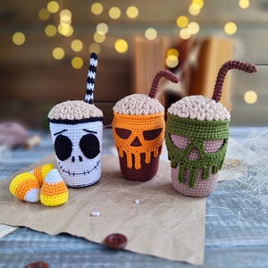 May include: Three crocheted Halloween-themed coffee cups with straws. The cups are decorated with a skull, a pumpkin, and a skeleton face. The cups are sitting on a brown surface with candy corn.