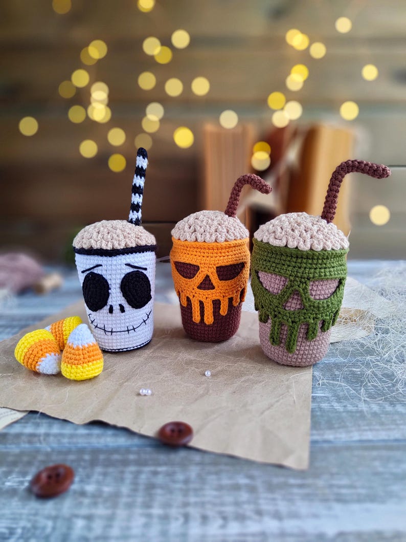 May include: Three crocheted Halloween-themed coffee cups with straws. The cups are decorated with a skull, a pumpkin, and a skeleton face. The cups are sitting on a brown surface with candy corn.