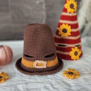 May include: A brown crocheted Pilgrim hat with an orange band and buckle, next to a red and white striped cone-shaped decoration adorned with yellow sunflowers. Additional yellow sunflower accents are scattered around.