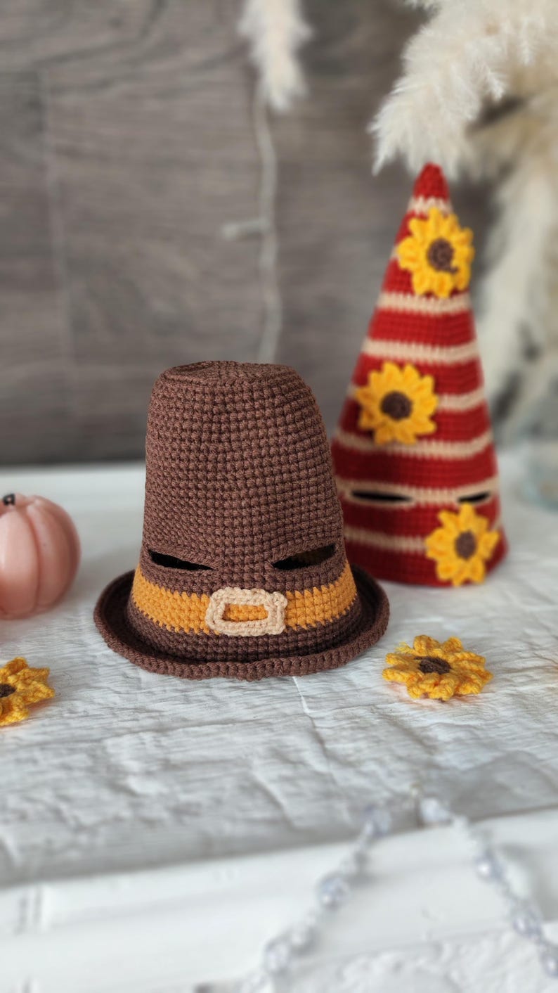 May include: A brown crocheted Pilgrim hat with an orange band and buckle, next to a red and white striped cone-shaped decoration adorned with yellow sunflowers. Additional yellow sunflower accents are scattered around.