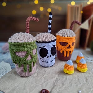 May include: Three crocheted Halloween-themed cups with different faces and straws. The cups are sitting on a brown paper surface with three crocheted candy corn pieces.