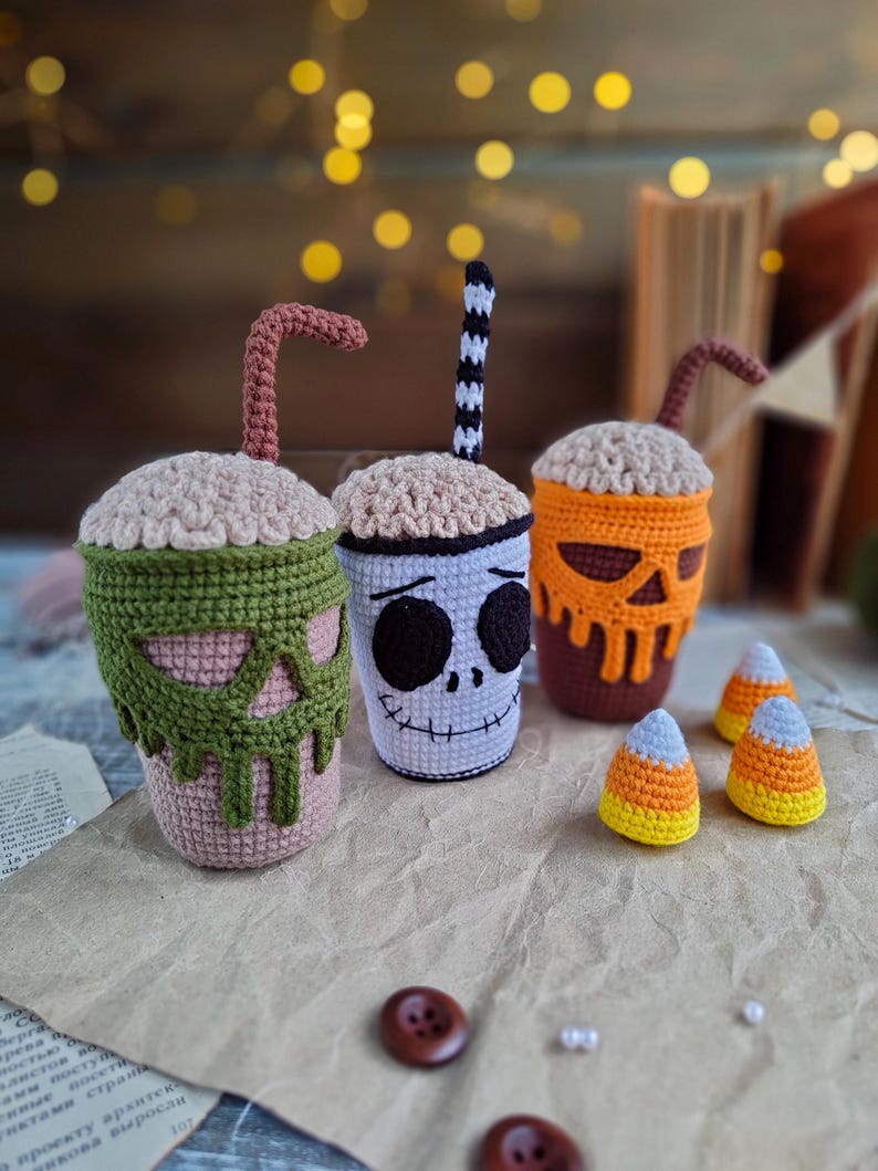 May include: Three crocheted Halloween-themed cups with different faces and straws. The cups are sitting on a brown paper surface with three crocheted candy corn pieces.