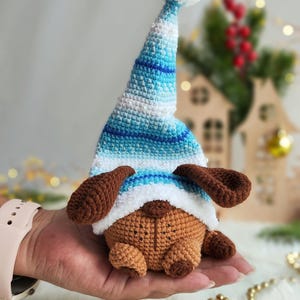 May include: A brown crocheted dog wearing a blue and white striped crocheted hat with a white pom-pom on top. The dog is sitting on a hand.