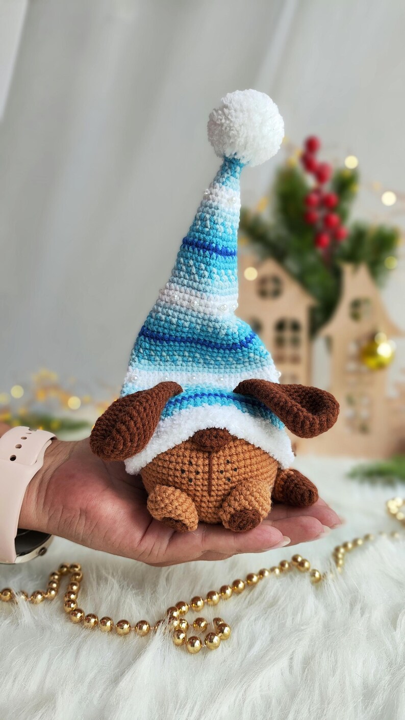 May include: A brown crocheted dog wearing a blue and white striped crocheted hat with a white pom-pom on top. The dog is sitting on a hand.