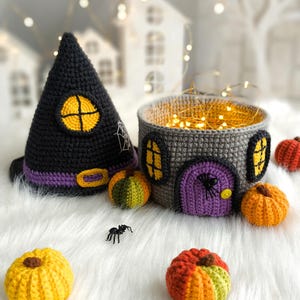 Op de afbeelding: Gehaakte Halloween-decoraties, waaronder een zwarte heksenhoed met een gele raam, een grijze huisvormige mand met een paarse deur en verschillende kleine pompoenen. Er is ook een kleine plastic spin aanwezig.