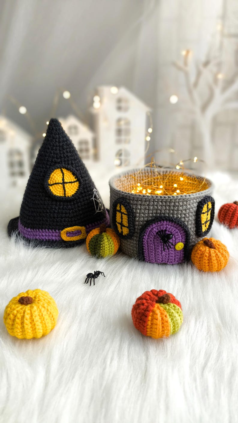 Op de afbeelding: Gehaakte Halloween-decoraties, waaronder een zwarte heksenhoed met een gele raam, een grijze huisvormige mand met een paarse deur en verschillende kleine pompoenen. Er is ook een kleine plastic spin aanwezig.