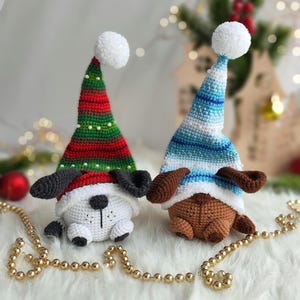 May include: Two crocheted dog ornaments wearing Christmas hats. One dog is white with black ears and a red and green hat. The other dog is brown with brown ears and a blue and white striped hat. The dogs are sitting on a white fluffy surface with gold beads.