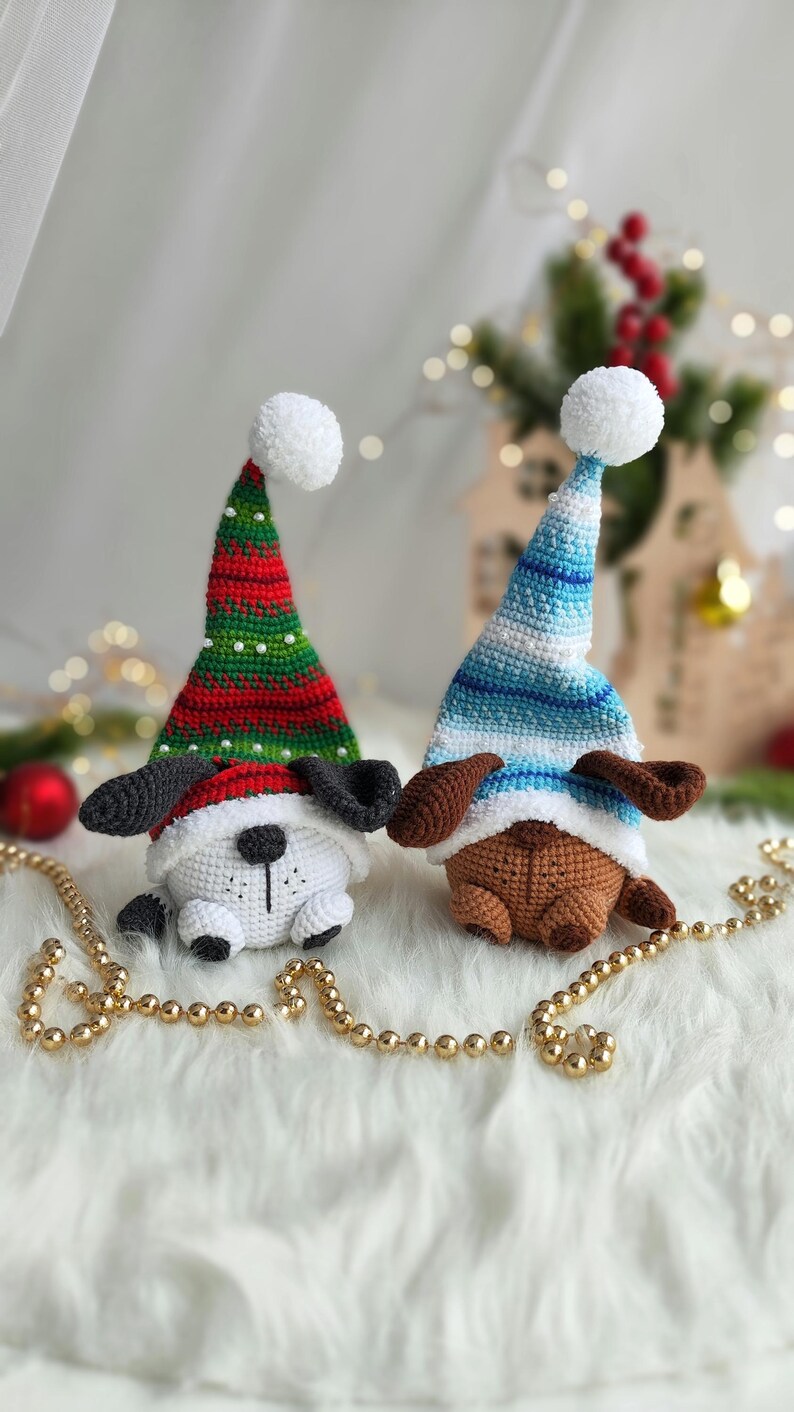 May include: Two crocheted dog ornaments wearing Christmas hats. One dog is white with black ears and a red and green hat. The other dog is brown with brown ears and a blue and white striped hat. The dogs are sitting on a white fluffy surface with gold beads.