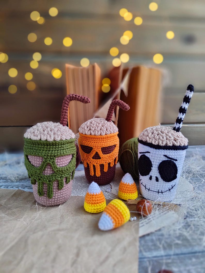 May include: Three crocheted Halloween-themed mugs with candy corn accents. The mugs are decorated with a skull, a pumpkin, and a skeleton face.
