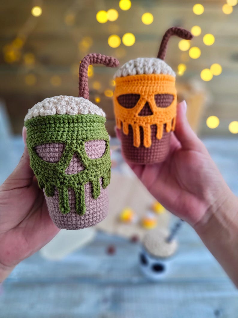 May include: Two crocheted Halloween-themed coffee cups with a skull design. One cup is green and the other is orange.