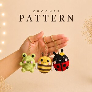 Puede incluir: Tres llaveros de ganchillo: una rana verde, una abeja amarilla y una mariquita roja. Cada llavero tiene un anillo dorado. La palabra "CROCHET PATTERN" se muestra en una fuente serif encima de los llaveros.