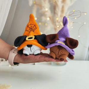 May include: Two crocheted Halloween dog toys wearing witch hats. One dog is white with black ears and an orange hat with a spiderweb design. The other dog is brown with purple hat.
