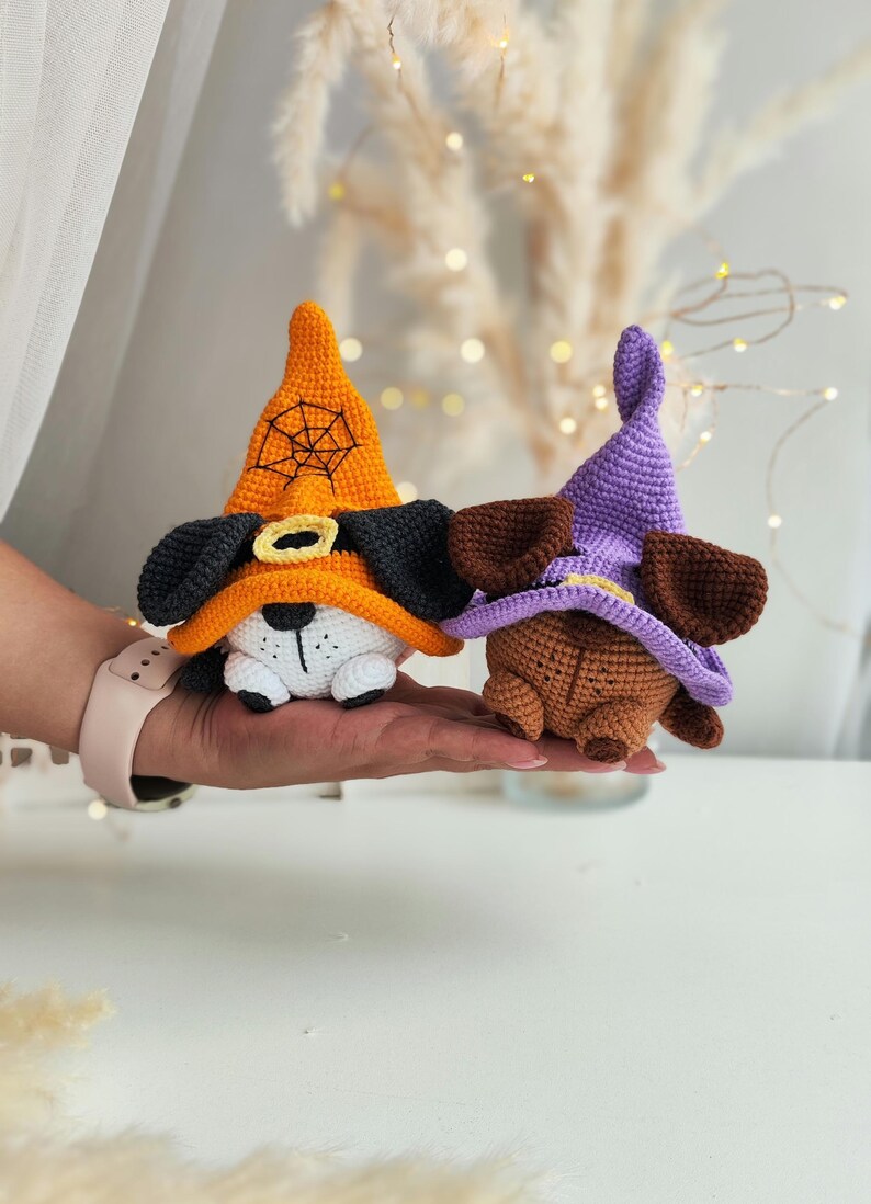 May include: Two crocheted Halloween dog toys wearing witch hats. One dog is white with black ears and an orange hat with a spiderweb design. The other dog is brown with purple hat.