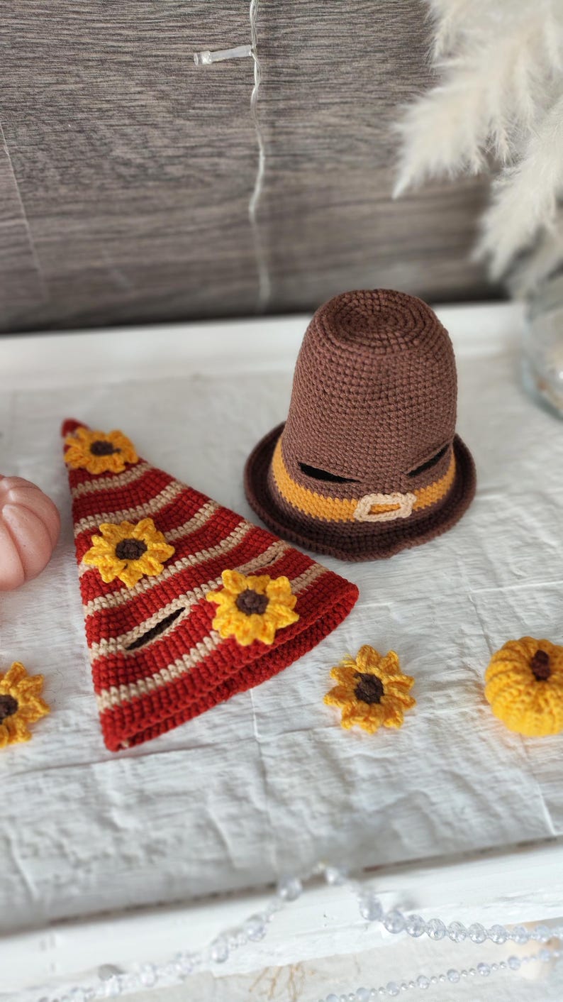 May include: Crocheted Thanksgiving-themed decorations. Includes a brown pilgrim hat with a yellow band and buckle, a red and cream striped hat with yellow sunflowers, and additional sunflower and pumpkin accents. The items are handmade.