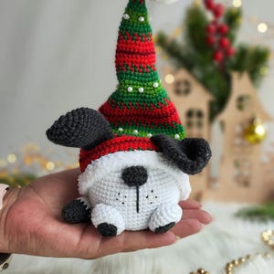 May include: A white crocheted dog wearing a red and green striped Santa hat with a white pom-pom on top. The dog is sitting on a hand.