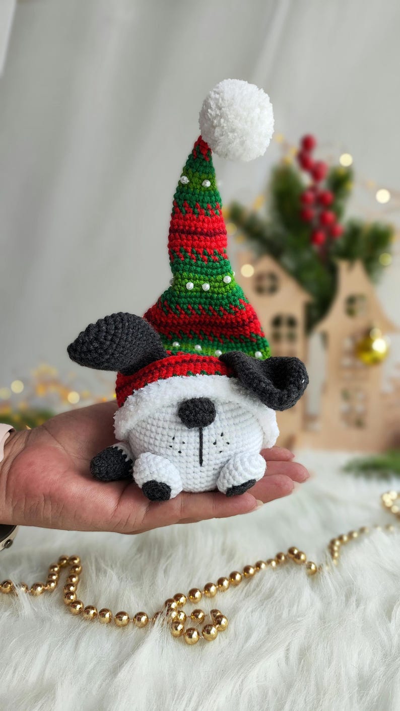 May include: A white crocheted dog wearing a red and green striped Santa hat with a white pom-pom on top. The dog is sitting on a hand.