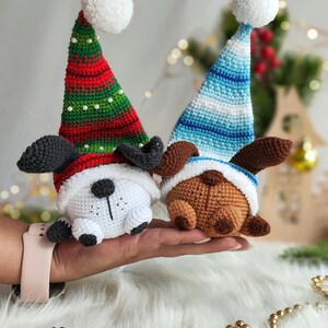 May include: Two crocheted dog ornaments wearing Santa hats. One dog is white with black ears and nose, the other is brown with black ears and nose. Both dogs are wearing white pom-pom hats with red and green stripes or blue and white stripes.