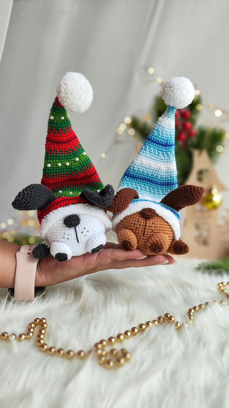 May include: Two crocheted dog ornaments wearing Santa hats. One dog is white with black ears and nose, the other is brown with black ears and nose. Both dogs are wearing white pom-pom hats with red and green stripes or blue and white stripes.