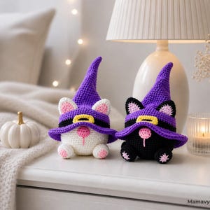 May include: Two crochet cat figurines wearing purple witch hats with black bands and yellow buckles. One cat is white with pink ears and paws, the other is black with pink ears and paws. The cats are Halloween-themed decorations.
