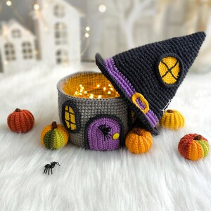 Op de afbeelding: Een gehaakt Halloween-huis met een heksenhoed, pompoenen en een spin. Het grijze huis heeft een paarse deur, gele ramen en is gevuld met fee&euml;rieke lichtjes. De zwarte heksenhoed heeft een gele gesp en een gele cirkel.