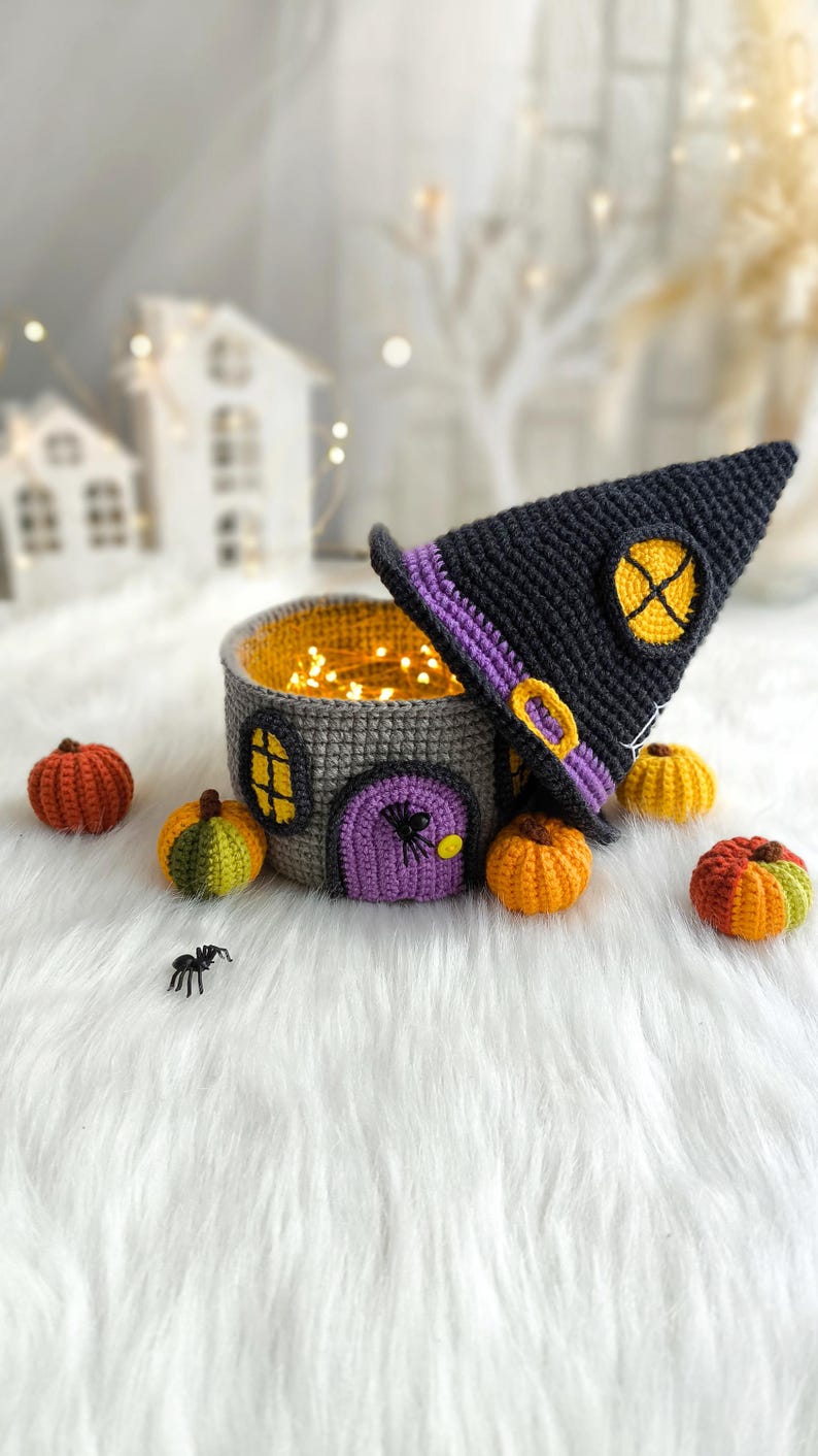 Op de afbeelding: Een gehaakt Halloween-huis met een heksenhoed, pompoenen en een spin. Het grijze huis heeft een paarse deur, gele ramen en is gevuld met fee&euml;rieke lichtjes. De zwarte heksenhoed heeft een gele gesp en een gele cirkel.