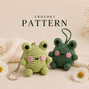 Pode incluir: Dois chaveiros de crochê de sapo, um verde claro e outro verde escuro, com olhos pretos e bochechas rosadas. Os chaveiros estão ao lado de flores brancas e das palavras "CROCHET PATTERN".