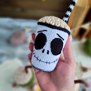 May include: A crocheted white cup with a black and white striped straw. The cup has a black stitched-on face with black eyes and a stitched-on smile. The cup is a Halloween-themed character.