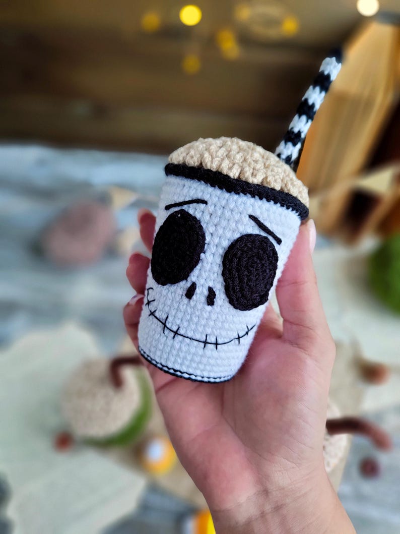 May include: A crocheted white cup with a black and white striped straw. The cup has a black stitched-on face with black eyes and a stitched-on smile. The cup is a Halloween-themed character.