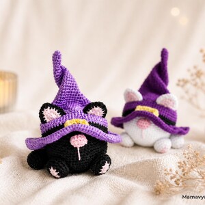 May include: Two crocheted cat gnomes wearing purple witch hats with yellow bands. One is black with pink accents, the other is white. The gnomes are on a cream-colored surface, with a lit candle and small pumpkin in the background. The text "MamavyazCrochet" is visible.