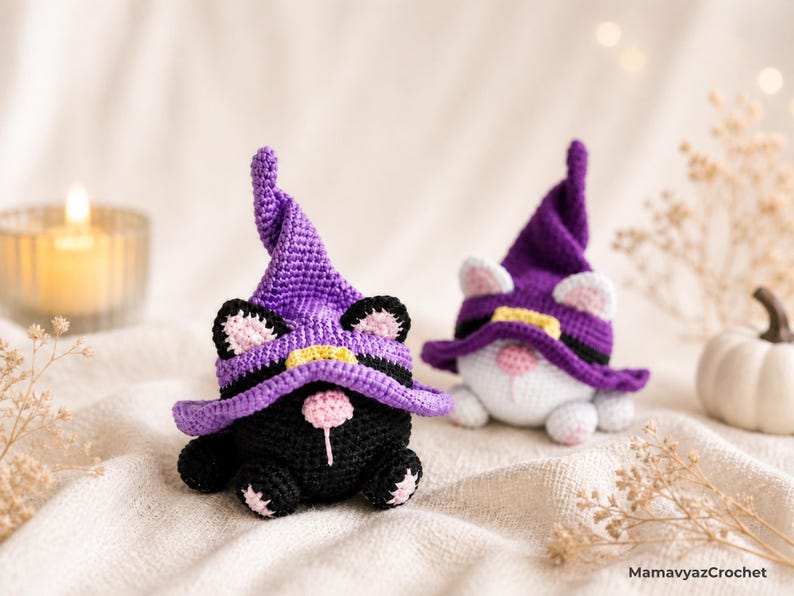 May include: Two crocheted cat gnomes wearing purple witch hats with yellow bands. One is black with pink accents, the other is white. The gnomes are on a cream-colored surface, with a lit candle and small pumpkin in the background. The text "MamavyazCrochet" is visible.