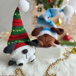 May include: Two crocheted dog ornaments wearing festive hats. One dog is wearing a red and green Santa hat with a white pom-pom, and the other is wearing a blue and white striped hat with a white pom-pom.
