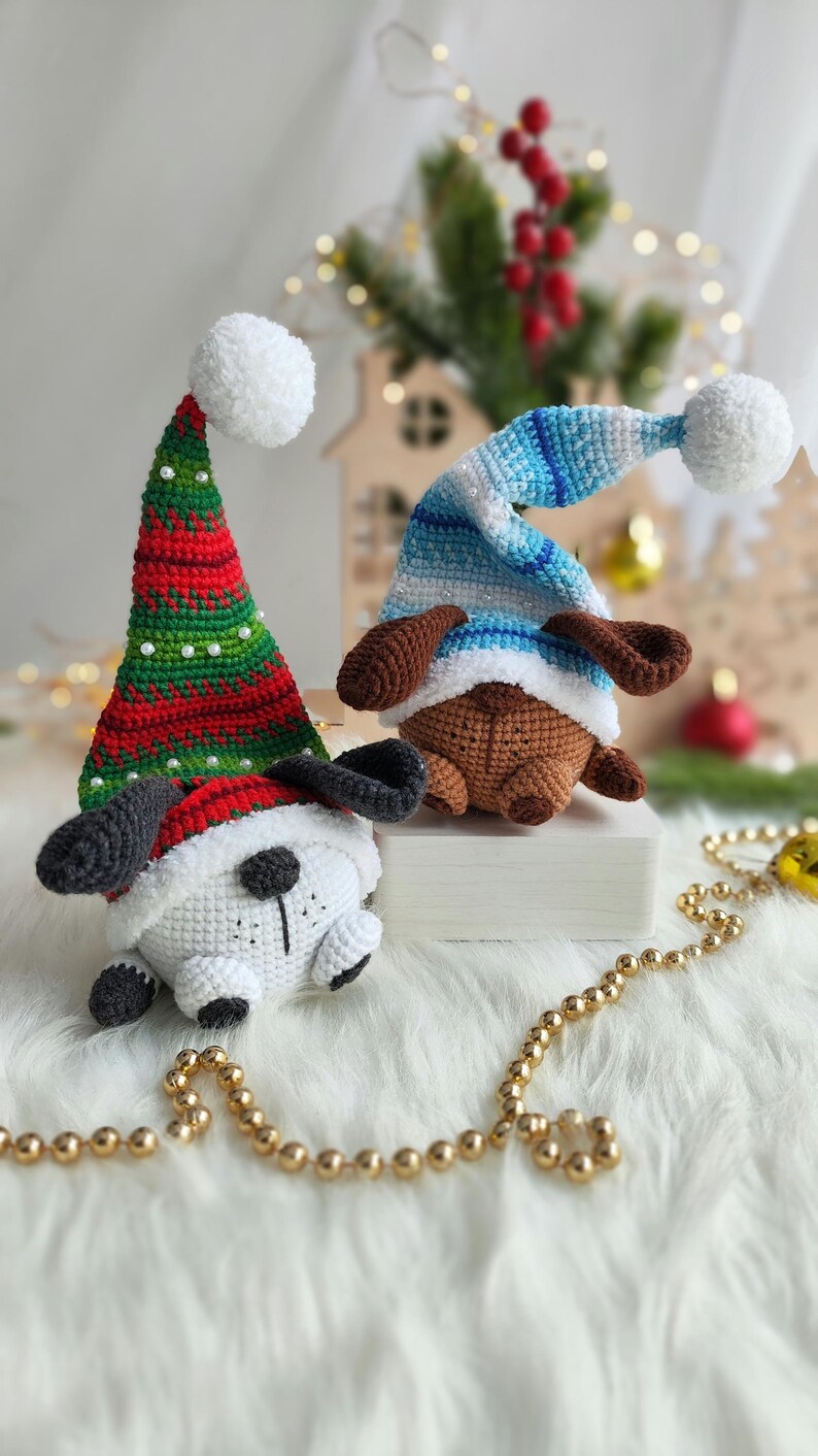 May include: Two crocheted dog ornaments wearing festive hats. One dog is wearing a red and green Santa hat with a white pom-pom, and the other is wearing a blue and white striped hat with a white pom-pom.
