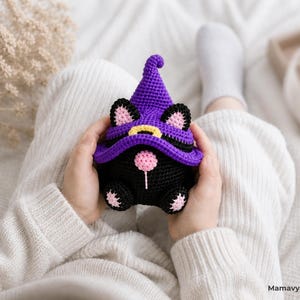 May include: A handmade black and purple crochet gnome with cat ears and a witch hat. The gnome has a pink nose and paws, and a yellow buckle on the hat. The gnome is held in someone's hands.