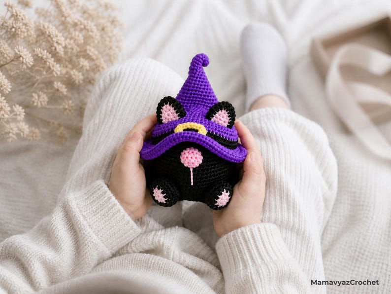 May include: A handmade black and purple crochet gnome with cat ears and a witch hat. The gnome has a pink nose and paws, and a yellow buckle on the hat. The gnome is held in someone's hands.