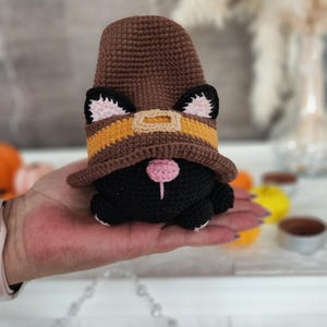 May include: A handmade crochet gnome with a black body, wearing a brown hat with a yellow and orange band. The gnome has black ears with pink inner ears and a pink nose. The gnome is being held in a person's hand.