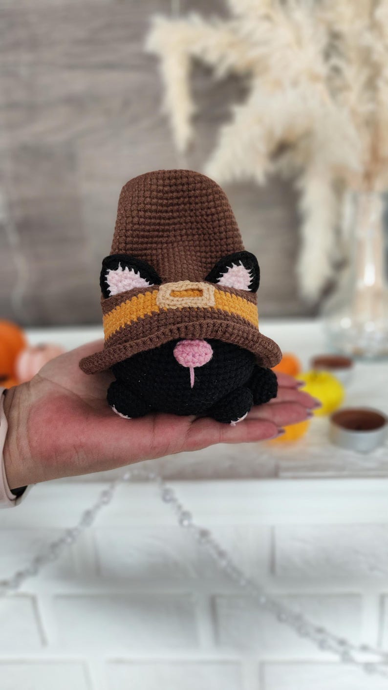 May include: A handmade crochet gnome with a black body, wearing a brown hat with a yellow and orange band. The gnome has black ears with pink inner ears and a pink nose. The gnome is being held in a person's hand.