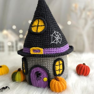Op de afbeelding: Een gehaakt Halloween-huis met een zwart heksenhoeddak, grijze muren en een paarse deur. De hoed heeft een gele raam en een wit spinnenweb. Kleine gehaakte pompoenen in oranje, geel en groen omringen het huis.