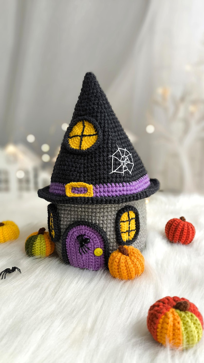 Op de afbeelding: Een gehaakt Halloween-huis met een zwart heksenhoeddak, grijze muren en een paarse deur. De hoed heeft een gele raam en een wit spinnenweb. Kleine gehaakte pompoenen in oranje, geel en groen omringen het huis.