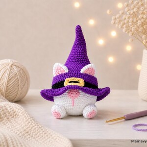May include: A handmade crochet gnome with a purple witch hat, black belt, and white body. The gnome has pink accents on its ears, nose, and feet. A ball of yarn, a crochet hook, and a vase with dried flowers are in the background.