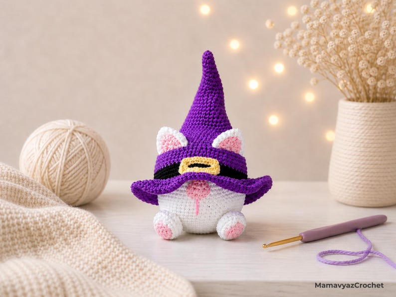 May include: A handmade crochet gnome with a purple witch hat, black belt, and white body. The gnome has pink accents on its ears, nose, and feet. A ball of yarn, a crochet hook, and a vase with dried flowers are in the background.