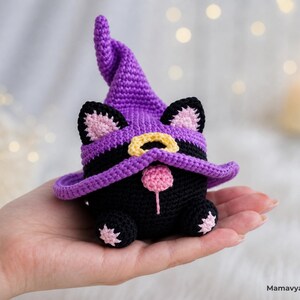 May include: A handmade black crochet cat wearing a purple witch hat with a yellow buckle and pink accents. The cat has pink ears and a pink nose. The cat is a Halloween or seasonal decoration.
