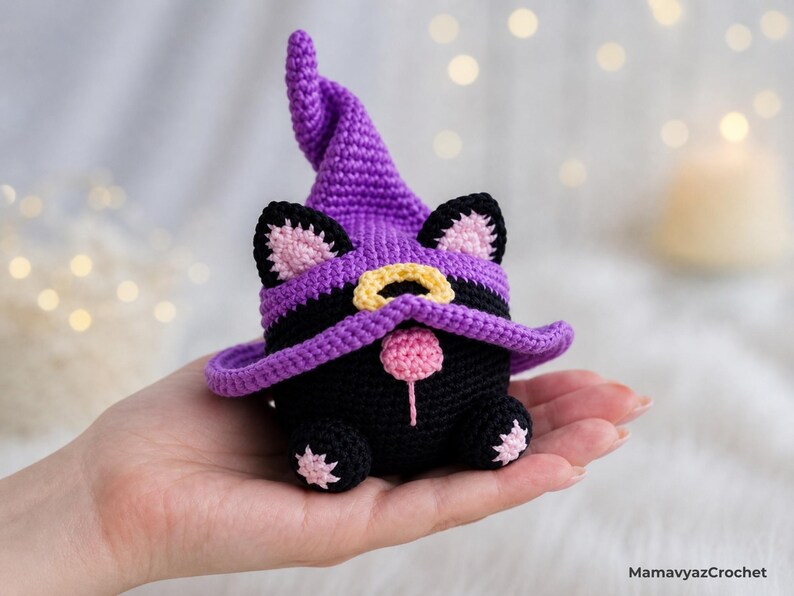 May include: A handmade black crochet cat wearing a purple witch hat with a yellow buckle and pink accents. The cat has pink ears and a pink nose. The cat is a Halloween or seasonal decoration.