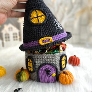 Op de afbeelding: Een gehaakte Halloween snoephouder in de vorm van een heksen huis. De zwarte hoed heeft een gele raam en een spinnenweb. Het grijze huis heeft een paarse deur en gele ramen. Miniatuur pompoenen en een spin zijn in de buurt.