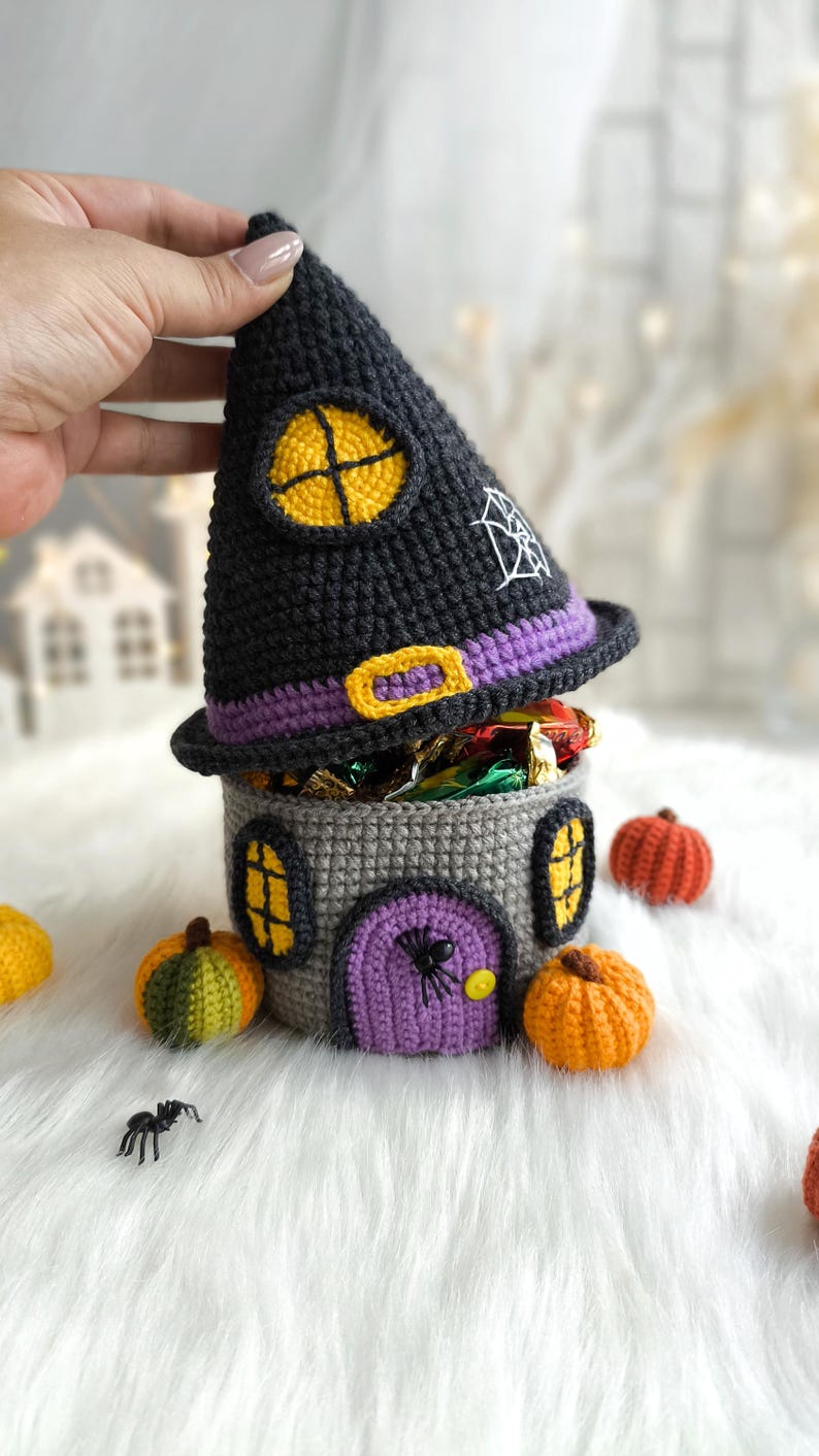 Op de afbeelding: Een gehaakte Halloween snoephouder in de vorm van een heksen huis. De zwarte hoed heeft een gele raam en een spinnenweb. Het grijze huis heeft een paarse deur en gele ramen. Miniatuur pompoenen en een spin zijn in de buurt.