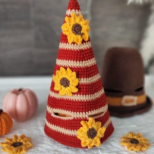 May include: A handmade crochet cone-shaped decoration with red and cream stripes, adorned with yellow and brown sunflowers. A brown crochet hat with a tan band is in the background. Other crochet pumpkins and sunflowers are also visible.